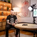 Фотография мастера тату, сидящего в кресле и дающего интервью. На заднем плане видна татуировка на стене и оборудование для татуировки.