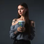 Фотография девушки с длинными темными волосами, держащей стакан кофе с изображением Земли. На ее руках видны детализированные татуировки.