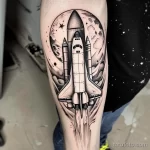 Фото тату: детализированная черно-белая татуировка космического шаттла на руке мужчины. Шаттл на фоне луны и звезд.