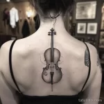 Фото тату виолончели на женской спине. Черно-белый рисунок татуировки с детальной проработкой корпуса инструмента и грифа.