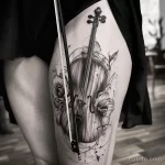 Фото тату виолончели на бедре в стиле blackwork с добавлением роз. Черно-белый рисунок с детальной проработкой и брызгами.