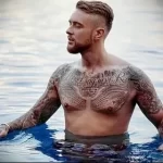 Фотография мужчины с татуировками на груди и руках, стоящего в воде, демонстрирующая сложные рисунки татуировки.