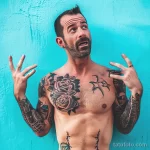 Фотография мужчины с татуировками на груди, руках и плечах. Татуировки выполнены в стиле реализм и графика.
