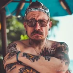Фотография мужчины в бандане и очках с татуировками в стиле олд скул на руках и груди. Татуировки выполнены в ярких цветах с четкими контурами.