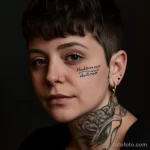Фото татуировки на лице и шее женщины. Изображена надпись, выполненная в черном цвете. Женщина с короткими волосами и пирсингом.