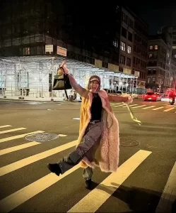 Фотография девушки, танцующей на пешеходном переходе в Нью-Йорке ночью. Она одета в длинное пальто и брюки.