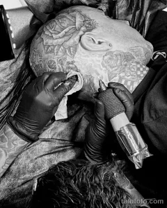 Фотография процесса нанесения татуировки на голову мужчины. Виден мастер тату, работающий с тату-машинкой и использующий салфетку.