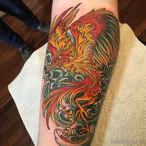 Фотография татуировки феникса на руке, выполненной в ярких оранжевых и желтых тонах. Детализированное изображение птицы в полете на фоне волн.