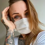 Фотография молодой женщины с татуировкой в виде крыла под глазом и яркими татуировками на руке, носящей медицинскую маску.