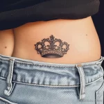 Фото тату с изображением короны на пояснице. Черно-белый рисунок с детализированными узорами и изящной формой. Подчеркивает грацию и уверенность.