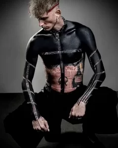 Фото тату Machine Gun Kelly, покрывающего тело черными татуировками с элементами граффити и надписями. Вид спереди, демонстрирующий масштаб рисунков.