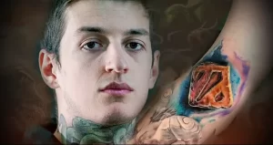 Фото тату с изображением логотипа Dota 2 на предплечье мужчины. Яркий рисунок татуировки выполнен в акварельной технике с использованием синих и красных оттенков.