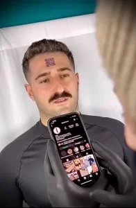 Фото тату: Мужчина с татуировкой QR-кода на лбу, демонстрирующий ее с помощью смартфона. Черные перчатки мастера.