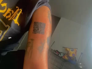 Фотография татуировки в виде QR-кода на предплечье мужчины. Рядом видны другие, менее четкие татуировки.