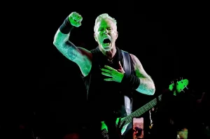 Фотография Джеймса Хетфилда, вокалиста Metallica, во время концерта. Видны татуировки на его руках и плечах. Энергичная и динамичная сцена.