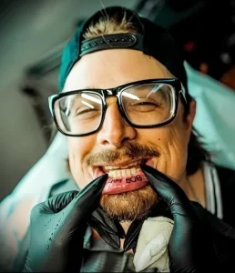 Фотография мужчины с татуировкой надписи "JIM BOB" на внутренней стороне губы. Мужчина широко улыбается, демонстрируя татуировку.