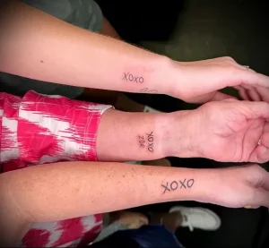 Фотография двух рук с парными татуировками 'XOXO'. Татуировки выполнены простым шрифтом черным цветом на внутренней стороне предплечья.