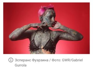 Фото тату: женщина с полностью покрытым татуировками телом, включая лицо и шею, позирует на красном фоне. Татуировки выполнены в темных тонах с яркими акцентами.