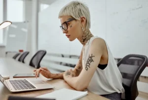 Девушка с татуировками на руках и шее работает за ноутбуком в офисе. Современный интерьер и деловая обстановка.