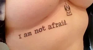 Фото минималистичной татуировки на груди девушки. Надпись 'I am not afraid' и небольшой символ.