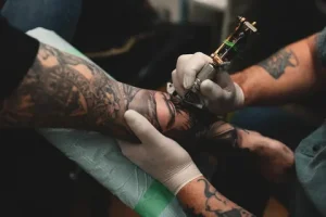 Фото тату процесса нанесения татуировки на руку. Мастер в перчатках работает тату-машинкой, а ассистент держит кожу.