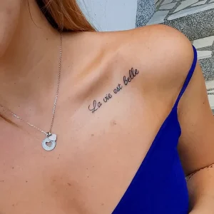 Изящная татуировка надписью «La vie est belle» на ключице девушки. Черный контур, тонкий шрифт, минималистичный дизайн.