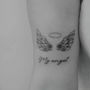 Миниатюрная татуировка ангельских крыльев с тонким нимбом и надписью "My angel", выполненная в графическом стиле на боку.
