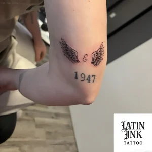 Татуировка ангельских крыльев с буквой 'Е' и числом '1947' на предплечье