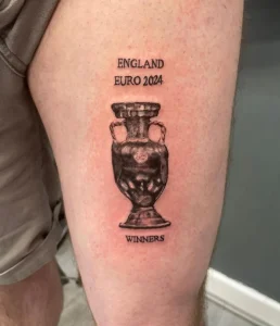 Фото тату на бедре с изображением кубка Евро 2024 и надписью 'ENGLAND EURO 2024 WINNERS'. Черно-белый рисунок татуировки в реалистичном стиле.