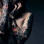 Фотография девушки с темными татуировками на руках, плечах и спине. Татуировки выполнены в реалистичном стиле и изображают черепа, цветы и другие мрачные символы.