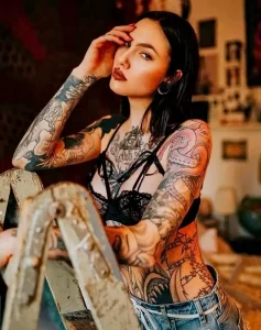 Фотография девушки с множеством ярких татуировок на руках, груди и боку. Татуировки выполнены в различных стилях и цветах.