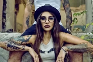 Фотография девушки в шляпе и очках с татуировками на руках. Она сидит на фоне кирпичной стены.