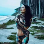 Фотография девушки с длинными темными волосами, покрытой татуировками на большей части тела. Она стоит на фоне моря и скал.