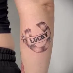 Фото тату с надписью 'Lucky' на ленте, стилизованной под старинную печать. Черно-белый рисунок татуировки, выполненный в графическом стиле на предплечье.