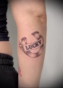 Фото тату с надписью 'Lucky' на ленте, стилизованной под старинную печать. Черно-белый рисунок татуировки, выполненный в графическом стиле на предплечье.