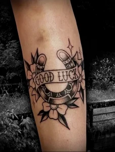 Фото тату на икре с изображением подковы, клевера и надписи 'GOOD LUCK' в стиле олд скул. Черно-белый рисунок татуировки.