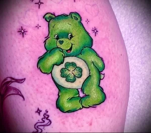 Фото яркой татуировки на руке: зеленый медвежонок Care Bear с четырехлистным клевером.