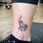Фото тату маленького слоненка на лодыжке. Черно-серый рисунок с красным камнем на голове слона. Детализированная проработка текстуры кожи.