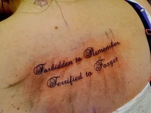 Фотография татуировки надписи "Forbidden to Remember, Terrified to Forget" на груди женщины. Рисунок выполнен в классическом шрифте.