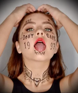 Фото тату: девушка с татуировками на лице, включая надпись 'I DON'T CARE', число '1997' и другие символы. Экстремальный и вызывающий рисунок.