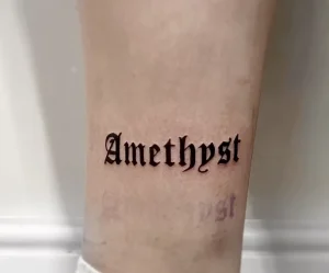 Фото минималистичной татуировки с надписью 'Amethyst' выполненной черными чернилами в готическом стиле на руке.