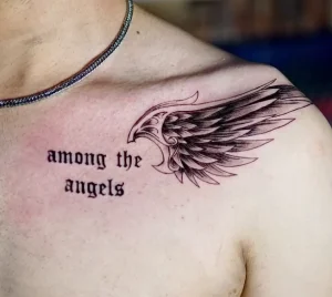 Фото тату: небольшое крыло и надпись 'among the angels' на груди, выполненные в графическом стиле. Минималистичный и трогательный дизайн.