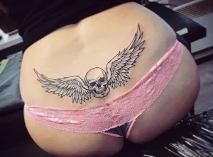 Фото тату черепа с крыльями на бедре. Черно-белый рисунок, крылья расположены по обе стороны от черепа, татуировка выглядит дерзко и загадочно.
