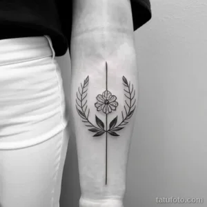 Фото тату на предплечье: черно-белый венок из листьев и цветка с вертикальной линией, создающий элегантный и минималистичный дизайн.