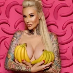 Фотография девушки с длинными светлыми волосами, держащей бананы перед лицом и демонстрирующей татуировки на руках и шее на фоне розовой стены с бананами.