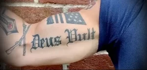 Крупный план татуировки на руке, изображающей американский флаг, надпись 'Deus Vult' и другие символы. Черно-белое изображение.