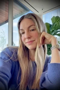 Женщина с длинными светлыми волосами, одетая в лавандовый свитер, смотрит в камеру с легкой улыбкой. На фоне окно и растения.