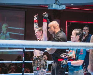 Фотография девушки-бойца MMA, празднующей победу после боя. Ее руку подняли вверх, и на ее теле видны татуировки.
