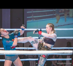 Фотография девушки-бойца MMA, наносящей мощный удар ногой в бою. На ее ноге и руках видны татуировки.