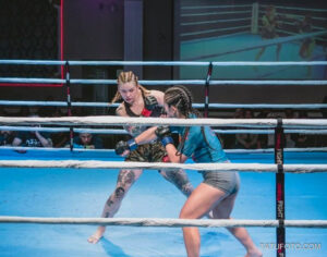 Фотография двух девушек-бойцов MMA в динамичном бою на ринге. Обе спортсменки имеют татуировки на руках и ногах.